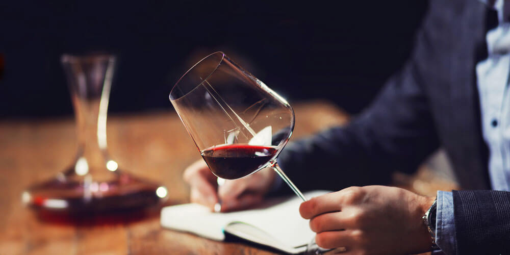 Mann sitzt mit einem Glas Rotwein in der Hand am Tisch auf dem ein gefüllter Decanter steht und schreibt in ein Buch
